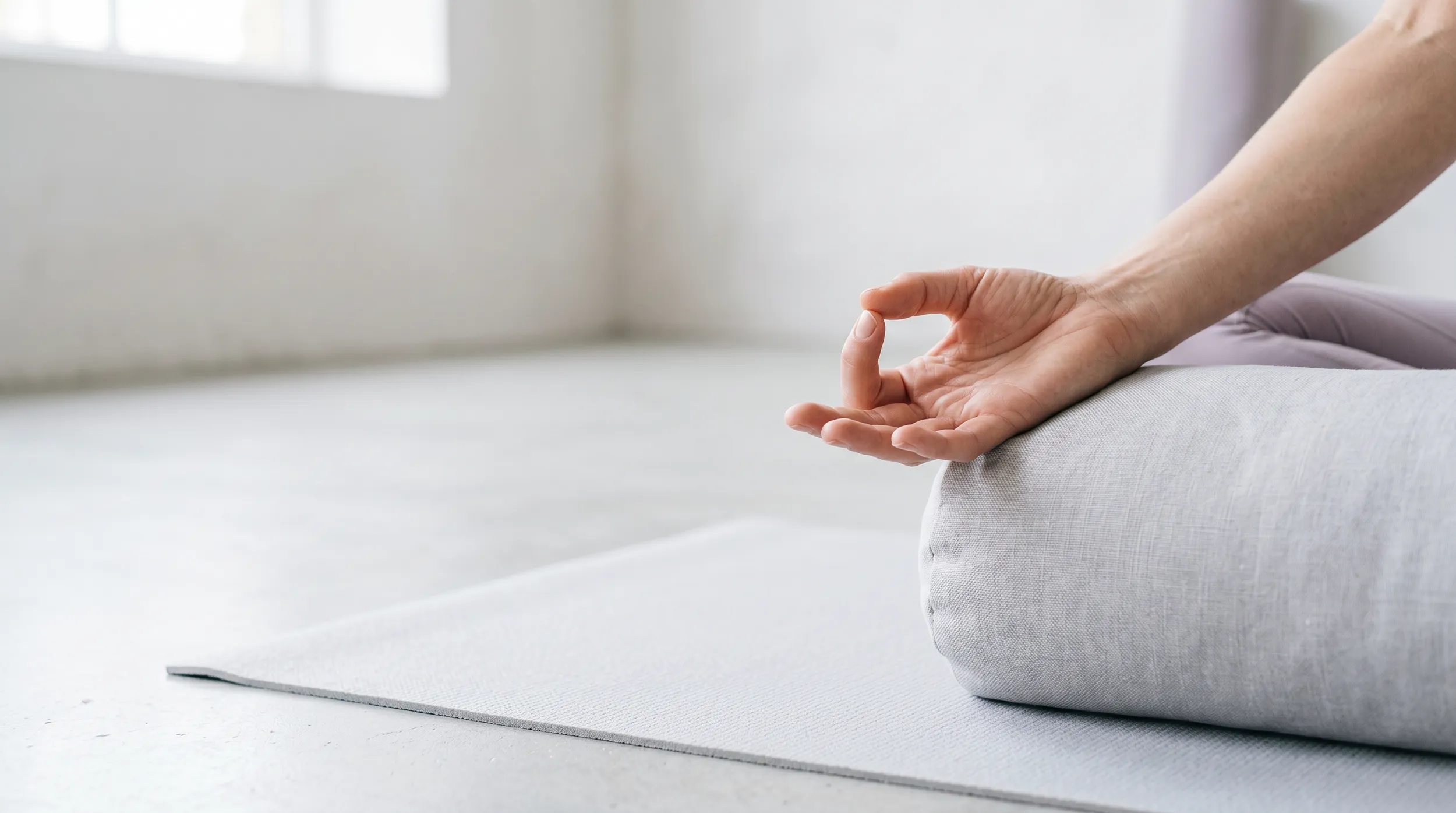 Hands resting in a meditation mudra on a cushion beside a yoga mat